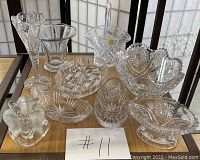 Photo showing all 11 vintage glass candy dishes arranged on a wooden table with window screens in the background. Various shapes including baskets, oval dishes, footed bowls, and scalloped edge dishes with starburst and lattice patterns.