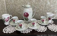 Wide shot showing tea pot, four small cups with saucers, and two larger mugs, all placed on a crocheted lace table runner.