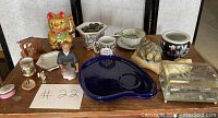 Photo of various vintage Asian collectible items including Maneki-neko cat figurine, elderly woman figurine, small wooden bench, ceramic cups, and dishes arranged on a wooden surface.