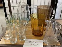 Lot of vintage pitchers and glasses displayed on a table showing a variety of clear and colored glassware with decorative etched and painted patterns.