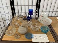 Various vintage glassware items arranged on a table including clear, white, blue pieces, bowls, jars, decanters, and lidded containers with floral painted details