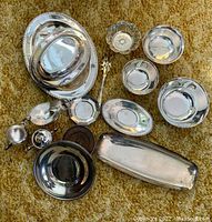 Overview of assorted vintage silver-plated serving trays, bowls, candle holders, and utensils on carpet.
