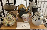 Photo showing eight vintage tea pots arranged on a wooden surface with a paper labeled #14. The tea pots vary in size, shape, and material including ceramic with floral designs and metal pots.