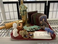 A tray with an assortment of vintage collectibles including a white bust, a brass candlestick, a rolling pin, a large face decoration, a blue cloisonné trinket box, and a group of embossed leather-look bookends in red and green.
