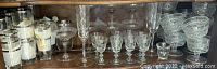 Wide display of multiple vintage cordial and glassware items arranged on a wooden shelf showing variety of sizes and etched patterns.
