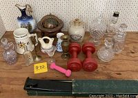 Wide view of the lot contents on wooden table showing glass decanters, ceramic items, dumbbells and metal and pottery collectibles.