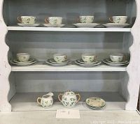 Full view of vintage shamrock tea set arranged on shelved white wooden hutch including 9 teacups with saucers, 4 additional saucers, a creamer, and sugar bowl with lid, all decorated with green shamrocks and gold trim.