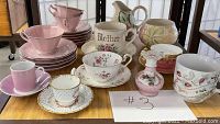 A display of multiple vintage collectible tea cups and saucers, along with pitchers and a small pink glass perfume bottle, arranged on a wooden shelf.