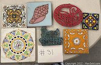 Seven vintage decorative trivets and tiles arranged on a table, showing a variety of colors and materials including ceramic tiles with floral and geometric designs and cast metal trivets shaped like a swan and a mythical creature.