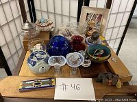 Overview of all items displayed on a wooden table with white folding screen background, showing assortment of glass vases, figurines, seashells, and decorative containers.