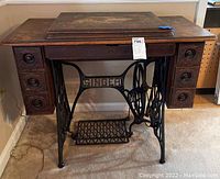 Front view showing wooden top with worn finish, cast iron base with treadle pedal, and six carved wooden drawers.
