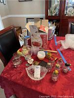 Full view of barware and kitchen items on red tablecloth including glass decanter, shot glasses, ice cream bowls, lidded jars, and wooden back scratchers.