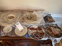 Full lot of seven decorative ceramic plates resting on bubble wrap on a wooden surface; includes Capodimonte style and Knowles/Reco collector plates.
