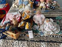 View of several porcelain and ceramic dolls placed on bedspread with colorful patterned blanket, including a clown doll and other dolls, alongside children's clothing.