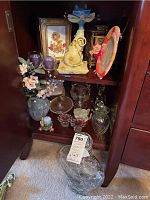 Shelf overview with ceramic vase and artificial flowers, glass candleholders with purple candles, cat figurine candle holder, picture frames, metal lantern, glass compote dish, small glass items, clear decanter and floor vase in front