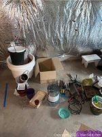 Overview shot showing large white and green planting pots stacked, various smaller pots, gardening tools, and accessories laid out on the floor against a reflective foil background.