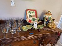 Full lot showing 12 zodiac glasses, fruit serving bowl, metal tray, three green nut bowls with stands, ceramic teapot, cookie jar, and two candlesticks on wooden sideboard.