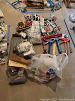 Photo shows a collection of household repair tools and parts spread on a concrete floor including caulk guns, small hardware boxes, rolls of tape, and an engraver tool with red handle.