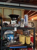 Full view of shelving unit with assorted small kitchen appliances and baking molds including Rival Hot Pot, wok, Chefman immersion blender, Gevalia coffee maker, baking pans, kitchen scale, and vintage Tupperware containers.