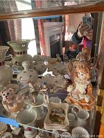 One shelf of porcelain Limoges figurines, trinket boxes, dishes and vases. Items arranged closely with reflected background showing fireplace and room. Items appear lightly dusty and require cleaning.