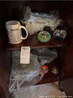 Shelf with white ceramic 'mom' mug, glass serving dishes wrapped in bubble wrap, and three glass paperweights