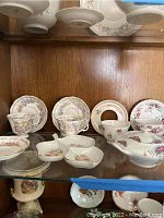 Shelf display of floral patterned china including cups and saucers, lidded serving dish, and sectioned serving bowl with gold trim on wooden shelving.