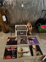 Photo of a white crate filled with vinyl records and a folding metal music stand behind it. Several album covers displayed in front on the floor.