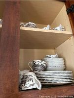 Stacked dinnerware and cups with black and white scenic transferware pattern inside a wooden cabinet.