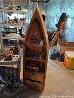 Full view of the wooden boat-shaped shelf standing in a basement. Shows three shelves and two drawers with some nautical decor on the shelves.