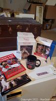 Photo shows the two Brad Lidge bobbleheads in original boxes, two Red Sox-themed tumblers in box, a black Boston University mug, and two books related to the Boston Red Sox including 'Faithful' and a commemorative book.