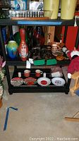 Multiple glass and porcelain vases, carved wood figurines, pottery, dinnerware, and plush toys on shelf and chairs in basement.