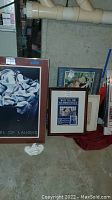 Photo showing five framed wall art pieces leaning against a basement wall including 'A Barrel of Laughs' poster and other prints.
