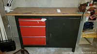 Front view of the metal workbench with wooden top showing the red drawers and black cabinet door, attached metal vice visible on the right side of the work surface.
