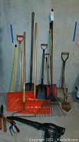 Photo showing various long-handled yard tools against a concrete basement wall including orange snow shovels, metal shovels, rakes with metal tines, and hand clippers in front with Remington blower lying horizontally.