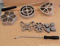 Photo showing eight Wagner Ware cast iron patty molds in various shapes including flower, circle, heart, butterfly, and small hearts, with two black-handled metal attachments for holding molds.