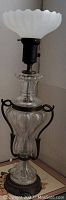 Full view of antique glass and metal lamp showing clear glass body with metal handles and white scalloped glass shade