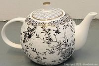 Side view showing intricate black floral and butterfly design on a white teapot with gold accents around lid.