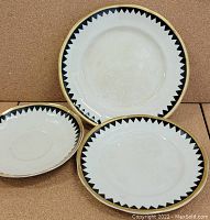 Three small plates with black triangular border pattern and gold edging, laid flat against a corkboard background.