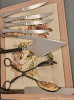 Top-down photo showing four dinner knives, a cake server, salad tongs, and a large serving spoon arranged on a floral print surface.