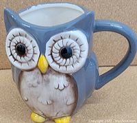 Close-up photo of a blue and brown owl-shaped ceramic mug with large eyes, yellow beak and feet, and handle on the right side, sitting on a flat surface.