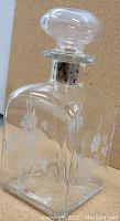 Close-up photo of one glass drink decanter showing floral etching and glass stopper with metal collar on neck.