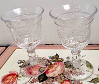 Two clear glass goblets with textured floral and geometric patterns on bowl, ornate stems, and round bases shown on a floral tray.