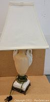 Beige ceramic lamp with swan motifs and white base, shown with its fabric lampshade and electrical cord with plug.