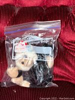 Front view of the Ty Beanie Baby plush monkey inside a clear resealable plastic storage bag on a red textured surface.