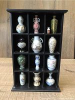 Full front photo of 13 miniature vases neatly arranged in a black wooden display case divided into 12 compartments showing their varied designs and colors.
