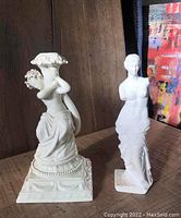 Two white ceramic woman figurines shown side by side on wooden surface, one larger signed Capo Di Monte with floral decoration, the other smaller classical style figure without arms.