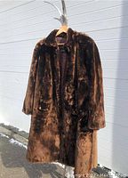 Front view of dark brown shaved beaver fur coat hanging on a hanger outside against a white wall, showing full front and detailing the collar, pockets and length.