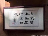 Framed Chinese calligraphy artwork with large black ink characters on white paper and accompanying smaller text and red seal stamp in lower left, under glass in dark wooden frame.