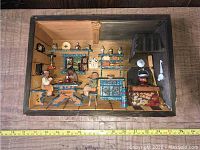Front view of the 3D painted wooden shadow box depicting a detailed farm/kitchen interior scene with furniture, two human figures, and various kitchen items.