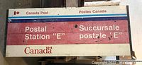 Photo of rectangular Canada Post metal sign with bilingual postal station "E" text, featuring red, blue, and white color scheme and Canada leaf logo in corner. Shows some wear and paint loss.